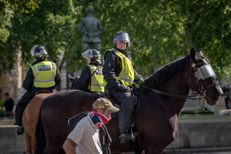 Police on horseback with helmets as a masked passerby in army cap passes by
